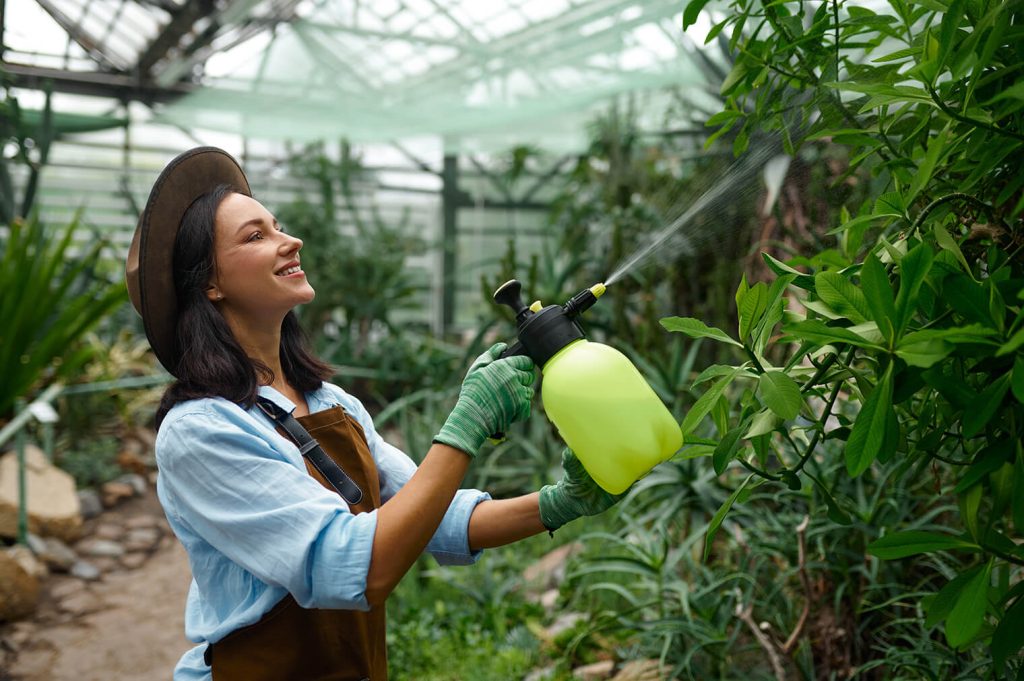 A smiling woman in a greenhouse sprays plants with a yellow spray bottle, wearing gloves and an apron. Green foliage surrounds her.